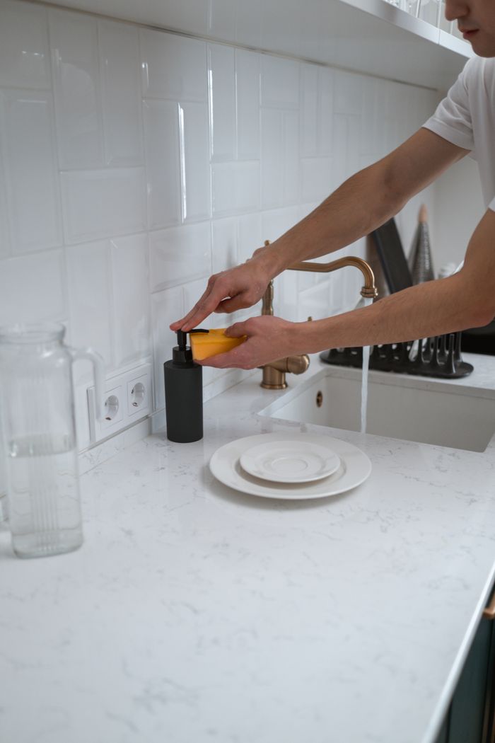 home-services-004 Close-up of hands washing dishes at a modern kitchen sink with white counter.